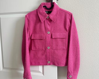 Könnte beinhalten: Eine rosafarbene Tweedjacke mit Knopfleiste und zwei Pattentaschen. Die Jacke hat eine verkürzte Länge und einen Kragen.
