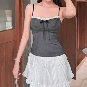 May include: A gray sleeveless top with a white lace trim and a black bow. The top is paired with a white skirt with a ruffled hem.