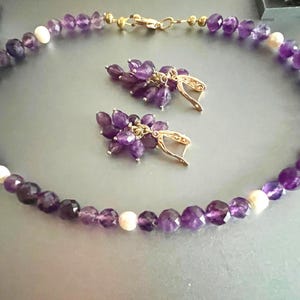 May include: A necklace and earring set featuring faceted purple amethyst beads interspersed with white pearls. The necklace and earrings have gold-colored metal accents. The earrings are clusters of amethyst beads.