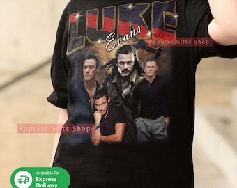 Puede incluir: Camiseta negra con un estampado gráfico de Luke Evans y el texto "LUKE Evans" en letras doradas con acentos rojos y azules. El gráfico también incluye un fondo de puesta de sol dorada.