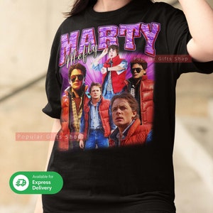 Puede incluir: Camiseta negra con un gr&aacute;fico morado y blanco de Marty McFly de la pel&iacute;cula Regreso al futuro. El gr&aacute;fico presenta m&uacute;ltiples im&aacute;genes de Marty McFly en diferentes escenas de la pel&iacute;cula. El texto "Marty McFly" est&aacute; escrito en letras moradas sobre el gr&aacute;fico. El texto "Popular Gifts Shop" est&aacute; escrito en letras blancas en el lado derecho del gr&aacute;fico.
