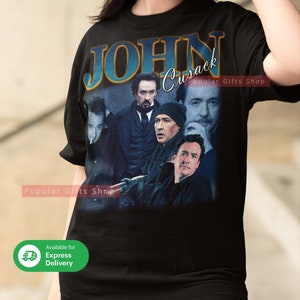 Op de afbeelding: Zwart T-shirt met een vintage-stijl grafische afbeelding van John Cusacks gezicht en de tekst "JOHN Cusack".