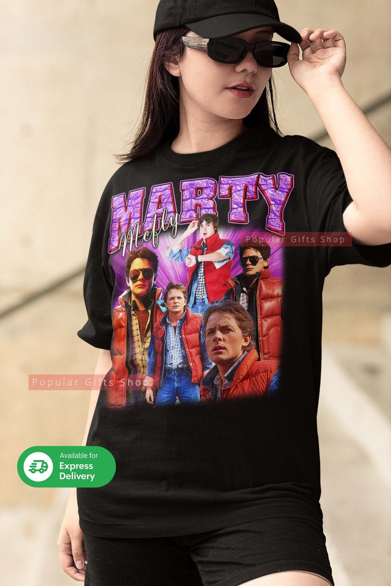 Puede incluir: Camiseta negra con un gr&aacute;fico morado y blanco de Marty McFly de la pel&iacute;cula Regreso al futuro. El gr&aacute;fico presenta m&uacute;ltiples im&aacute;genes de Marty McFly en diferentes escenas de la pel&iacute;cula. El texto "Marty McFly" est&aacute; escrito en letras moradas sobre el gr&aacute;fico. El texto "Popular Gifts Shop" est&aacute; escrito en letras blancas en el lado derecho del gr&aacute;fico.