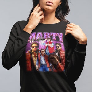 Puede incluir: Sudadera negra con un estampado gr&aacute;fico de Marty McFly y el elenco de Regreso al futuro. El estampado presenta un dise&ntilde;o morado y blanco con el texto "Marty McFly" y "Marty" en letras grandes.