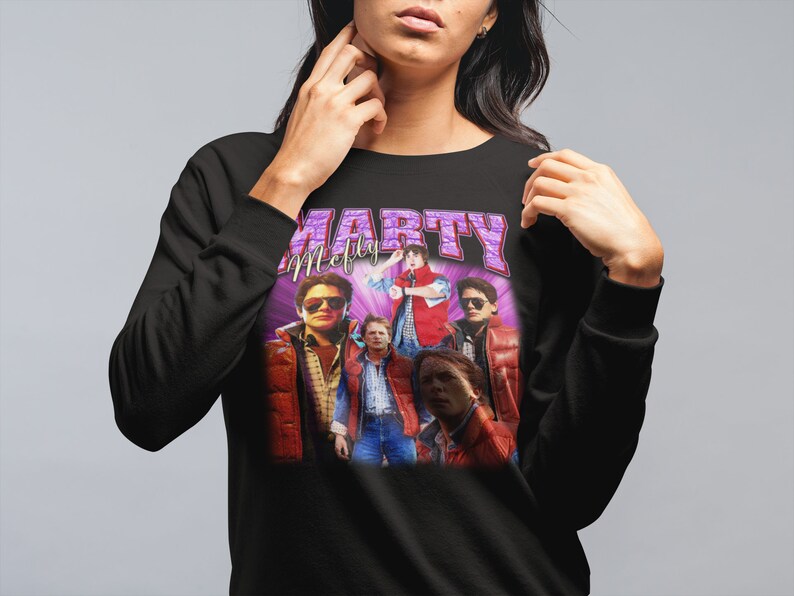 Puede incluir: Sudadera negra con un estampado gr&aacute;fico de Marty McFly y el elenco de Regreso al futuro. El estampado presenta un dise&ntilde;o morado y blanco con el texto "Marty McFly" y "Marty" en letras grandes.