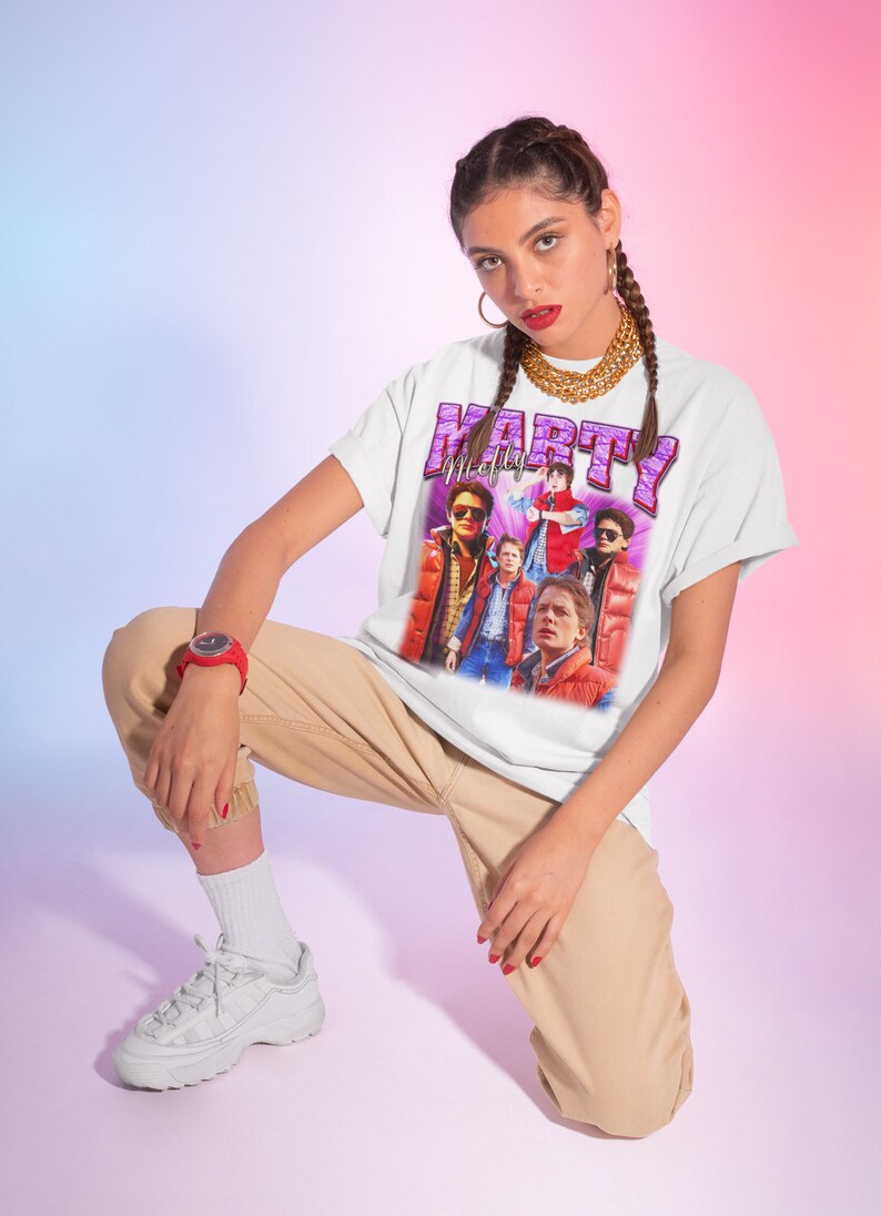 Puede incluir: Una camiseta blanca con un estampado gr&aacute;fico de un grupo de personas con chaquetas rojas y el texto "Marty McFly" en letras moradas.