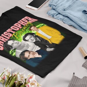 May include: Black t-shirt with a graphic print of a man's face with the text "CHRISTOPHER BRINLEY" in pink and yellow.
