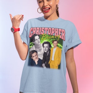 May include: Light blue t-shirt with a graphic print of a collage of four photos of Christopher Briney. The text "CHRISTOPHER Briney" is printed in pink letters above the photos.