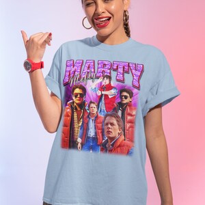 Puede incluir: Camiseta azul claro con un estampado gr&aacute;fico vintage de Marty McFly de la pel&iacute;cula Regreso al futuro. El gr&aacute;fico es un collage de diferentes im&aacute;genes de Marty McFly en varias escenas de la pel&iacute;cula. El texto "Marty McFly" est&aacute; impreso en letras moradas sobre el gr&aacute;fico.