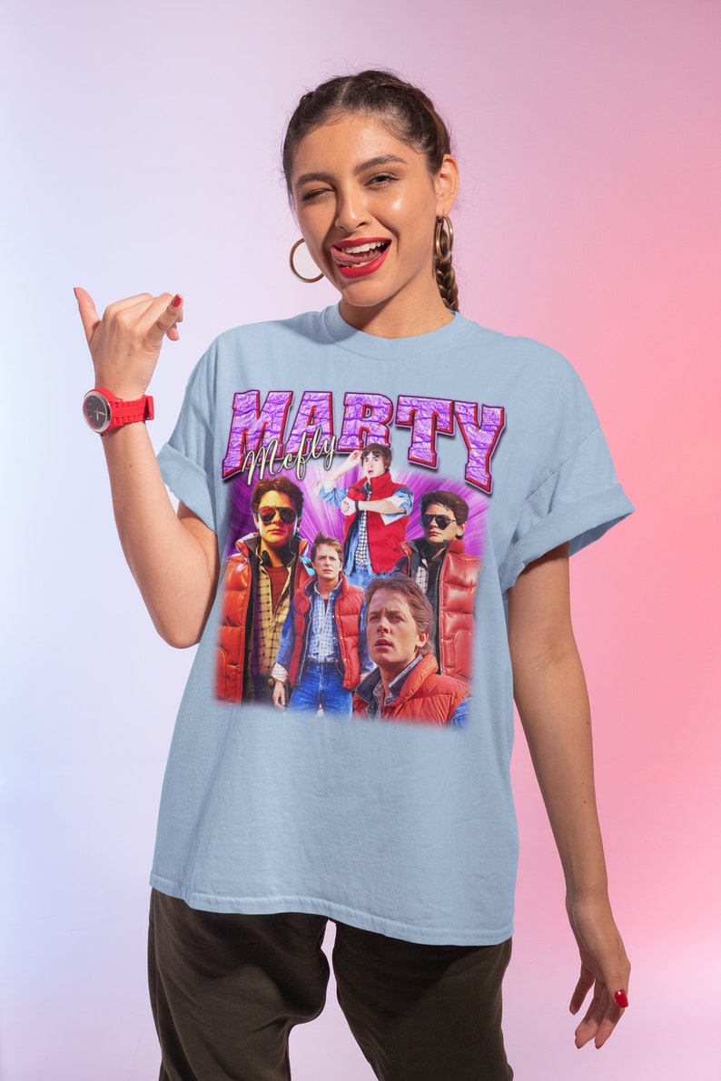Puede incluir: Camiseta azul claro con un estampado gr&aacute;fico vintage de Marty McFly de la pel&iacute;cula Regreso al futuro. El gr&aacute;fico es un collage de diferentes im&aacute;genes de Marty McFly en varias escenas de la pel&iacute;cula. El texto "Marty McFly" est&aacute; impreso en letras moradas sobre el gr&aacute;fico.