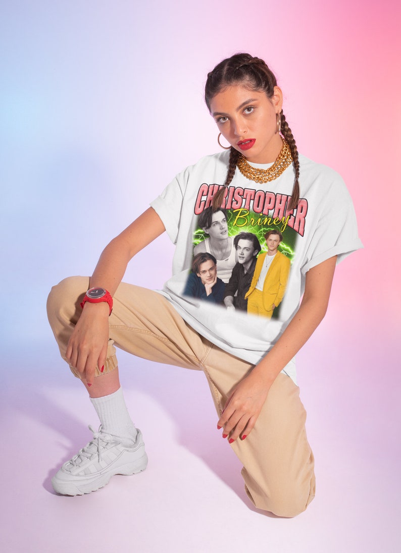 May include: A white t-shirt with a graphic print of a band called "Christopher Briney". The graphic features four men in a collage style. The shirt is being modeled by a person wearing khaki pants and white sneakers.