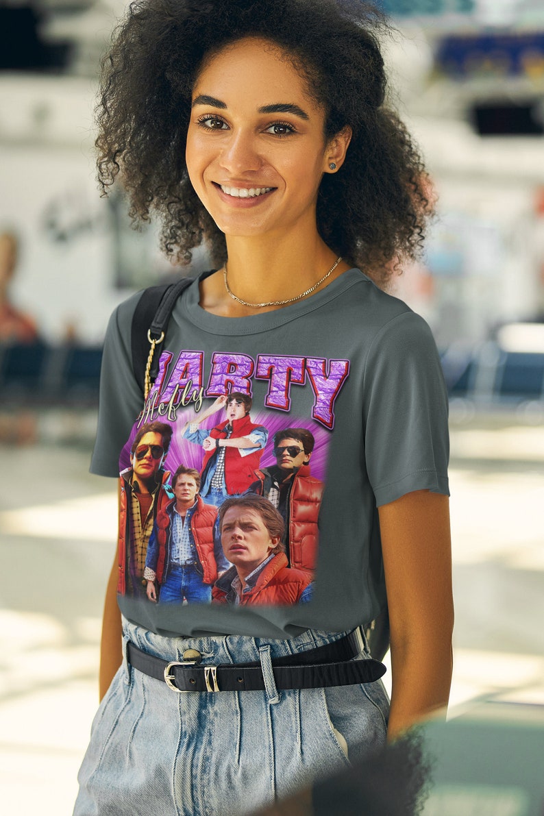 Puede incluir: Una camiseta gris con un estampado gr&aacute;fico de estilo vintage de Marty McFly de la pel&iacute;cula Regreso al futuro. El gr&aacute;fico presenta un texto "Marty" morado y un collage de im&aacute;genes de Marty McFly en diferentes escenas de la pel&iacute;cula.