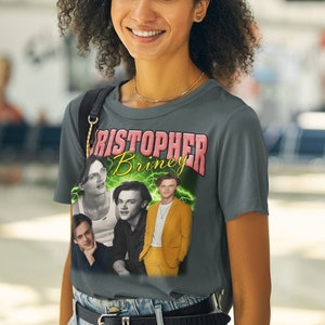 May include: A gray t-shirt with a graphic print of four men. The print features the text "Christopher Briney" in pink and green letters. The men are wearing different outfits, including a white tank top, a black shirt, a black shirt with a white collar, and a yellow suit.