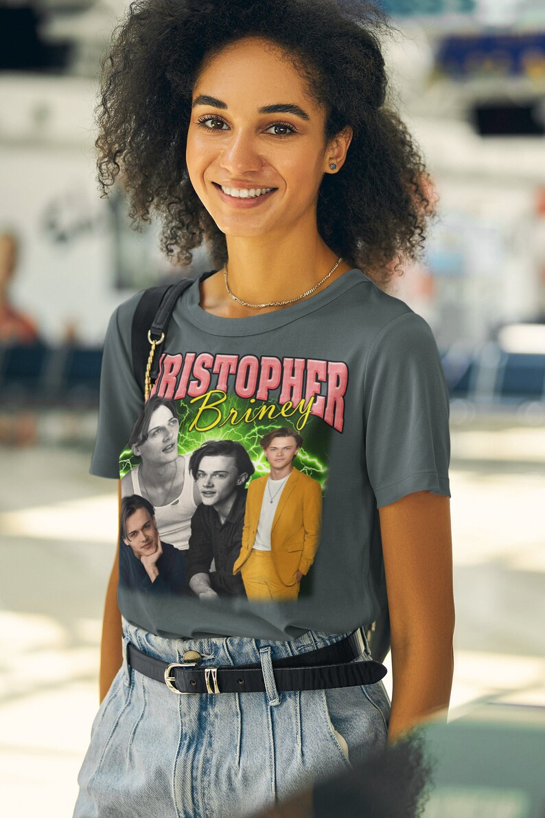 May include: A gray t-shirt with a graphic print of four men. The print features the text "Christopher Briney" in pink and green letters. The men are wearing different outfits, including a white tank top, a black shirt, a black shirt with a white collar, and a yellow suit.