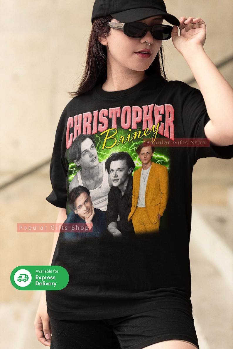 May include: Black t-shirt with a collage of images of Christopher Briney, a popular actor. The text "Christopher Briney" is printed in pink letters on the shirt. The text "Popular Gifts Shop" is printed in white letters on the shirt.