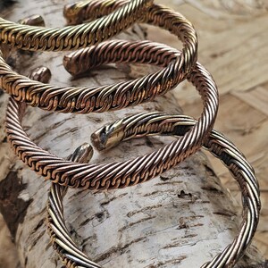 May include: Four textured metal cuff bracelets in shades of gold and copper are displayed. The bracelets have a twisted rope design and are arranged on a light-colored wooden surface. The bracelets are open-ended and appear to be adjustable.