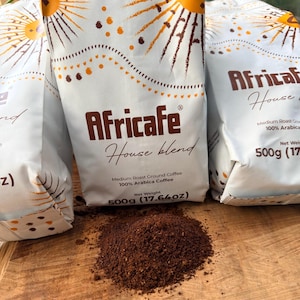 Op de afbeelding: Drie witte zakken Africafe House Blend medium gebrande gemalen koffie, elk met een gewicht van 500g. De verpakking heeft een zon-afbeelding met oranje en bruine stralen. Een hoopje donkerbruine gemalen koffie ligt voor de zakken.