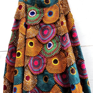 May include: A vibrant, tiered skirt with a colorful African print. The skirt features a repeating pattern of circular designs in shades of orange, teal, pink, and green. The skirt is hanging on a hanger against a white background.