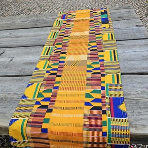 Peut inclure: Un chemin de table rectangulaire arborant un motif inspiré du Kente aux couleurs vives, avec des bandes géométriques jaunes, bleues, vertes, rouges et noires et des motifs tissés complexes disposés en bandes verticales sur le tissu.