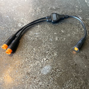Könnte beinhalten: Ein schwarzer elektrischer Stecker mit drei orangefarbenen Steckern an einem Ende und einem orangefarbenen Stecker am anderen Ende.