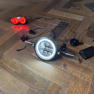 May include: Motorcycle headlight with a bright white halo and multiple LED lights. The image also shows a red taillight, a horn, and a black control box, all connected with wires. The items are on a wooden floor.
