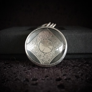 May include: A round, silver-toned pendant with a clear, domed glass center. The glass reveals an intricate, engraved design featuring geometric shapes and patterns. The pendant is suspended from a silver bail, set against a dark background.