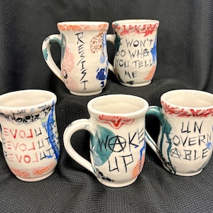 May include: Five handmade ceramic mugs with white, blue, red, and green accents. Each mug features unique hand-painted text, including "REVOLUTION," "WAKE UP," and "I WON'T DO WHAT YOU TELL ME." The mugs have curved handles and a rustic aesthetic.