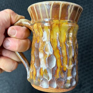 Puede incluir: Taza de cerámica hecha a mano con un patrón texturizado en forma de panal. La taza presenta un esmalte marrón, amarillo y blanco, con un borde ancho y un asa resistente. La taza está diseñada para bebidas calientes.