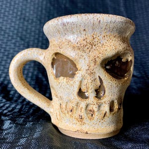 Pode incluir: Uma caneca de cerâmica em forma de caveira com esmalte marrom e branco. A caneca tem uma alça e uma abertura larga.