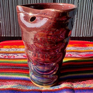 May include: A handmade ceramic vase with a rich, glossy burgundy glaze. The vase has a unique, textured design with a small hole near the top. It sits on a colorful, striped textile.