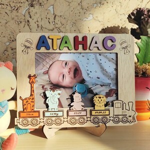 May include: Wooden photo frame with the name "ATAHAC" in colorful letters. The frame features a baby's photo and decorative animal train cars with birth details. A plush toy and a potted plant are also in the image.