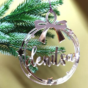 Puede incluir: Un adorno de Navidad de espejo de color oro rosa con la palabra "denitsa" escrita en cursiva dentro de un círculo con estrellas. El adorno tiene un lazo en la parte superior.