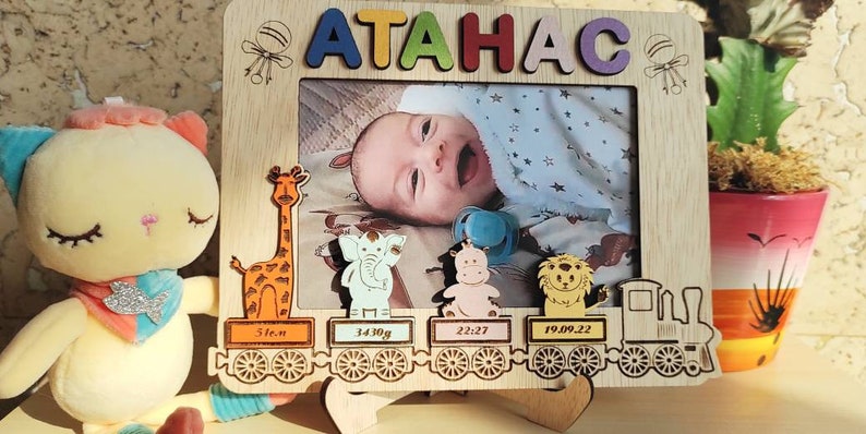 May include: Wooden photo frame with the name "ATAHAC" in colorful letters. The frame features a baby's photo, along with decorative animal cutouts and a train design. Includes baby's birth details: 31cm, 3430g, 22:27, and 19.09.22.