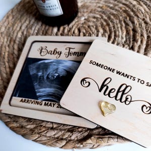 Op de afbeelding: Houten zwangerschapsaankondiging met een echografiebeeld en de tekst "Baby Tomm" en "Arriving May 20". De aankondiging bevat ook de tekst "Someone wants to say hello" en een gouden hartvormige bedel.