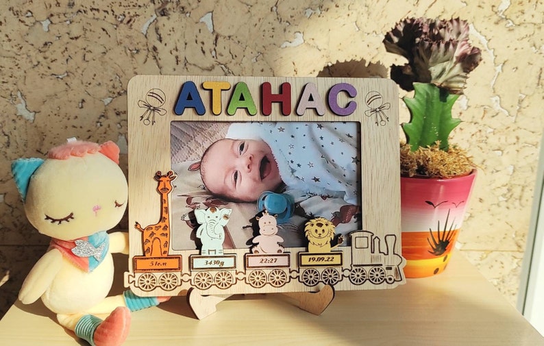 May include: Wooden photo frame with the name "ATAHAC" in colorful letters. The frame features a baby's photo and decorative animal train cars with birth details. A plush toy and a potted plant are also in the image.