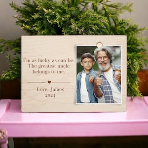 Puede incluir: Un marco de fotos de madera de color claro con una fotografía de un hombre y un niño haciendo una señal de pulgar hacia arriba. El marco tiene el texto "I'm as lucky as can be. The greatest uncle belongs to me. Love, James 2024."