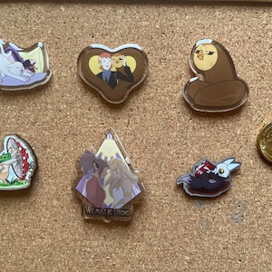May include: A selection of acrylic pins with cartoon illustrations. The pins depict a white cat, two figures in a heart, a brown owl, a mushroom with a frog, two figures with the text "WE MUST BE STRONG", and a bird-like creature. A 50-cent coin is also visible.