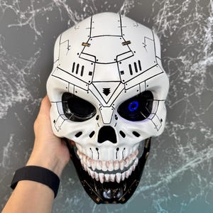 Può includere: Una maschera a teschio bianca con dettagli neri e un occhio blu brillante. La maschera ha un design meccanico con motivi geometrici e una mascella inferiore nera. La maschera è tenuta da una mano.