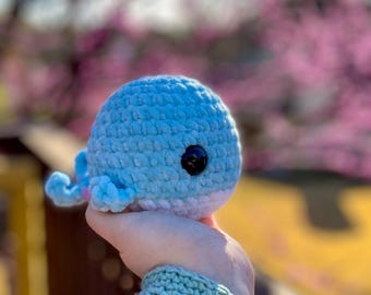 May include: A crocheted blue whale plush toy with a pink belly and black eyes. The whale is being held by a hand wearing a light green crocheted wrist warmer.