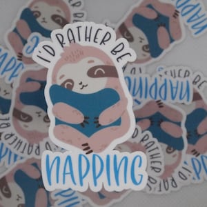 May include: A white sticker with a cartoon sloth holding a blue pillow. The sticker says "I'd rather be napping".