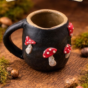 May include: A black ceramic mug with a handle, adorned with several red and white mushroom decorations. The mug has a beige rim and the text "Whimsical Mushroom Mug" on a banner.