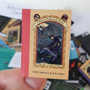 Peut inclure: Un livre de poche intitul&eacute; "L'Ascenseur de Remplacement" de la s&eacute;rie "Les D&eacute;sastreuses Aventures des Orphelins Baudelaire" par Lemony Snicket. La couverture pr&eacute;sente une illustration bleu fonc&eacute; d'une personne tombant dans un puits.