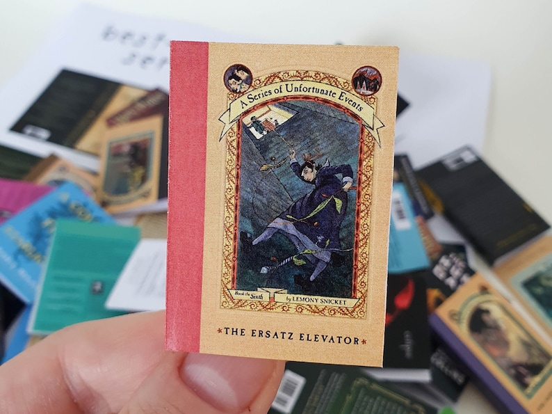 Peut inclure: Un livre de poche intitul&eacute; "L'Ascenseur de Remplacement" de la s&eacute;rie "Les D&eacute;sastreuses Aventures des Orphelins Baudelaire" par Lemony Snicket. La couverture pr&eacute;sente une illustration bleu fonc&eacute; d'une personne tombant dans un puits.