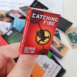 Peut inclure: Un livre de poche miniature avec une couverture rouge et des illustrations en noir et blanc. Le titre est "L'Embrasement" et l'auteur est Suzanne Collins. Le livre est le deuxi&egrave;me livre de la trilogie Hunger Games.
