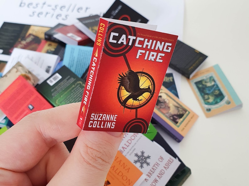 Peut inclure: Un livre de poche miniature avec une couverture rouge et des illustrations en noir et blanc. Le titre est "L'Embrasement" et l'auteur est Suzanne Collins. Le livre est le deuxi&egrave;me livre de la trilogie Hunger Games.