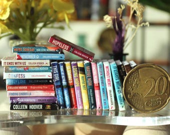 Puede incluir: Una colección de libros en miniatura de Colleen Hoover, incluyendo títulos como "Hopeless", "It Ends With Us" y "Maybe Now". Una moneda de 20 Euro Cent está junto a los libros. Los libros están apilados y alineados sobre una superficie transparente.