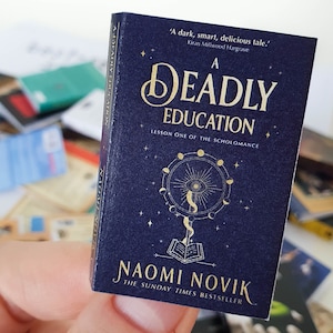 Peut inclure: Un livre bleu fonc&eacute; avec des lettres dor&eacute;es intitul&eacute; "A Deadly Education: Lesson One of the Scholomance" par Naomi Novik. La couverture du livre pr&eacute;sente une illustration dor&eacute;e d'un livre, d'un &oelig;il et d'un croissant de lune.