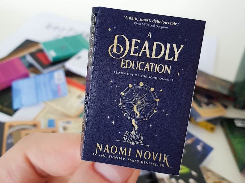 Peut inclure: Un livre bleu fonc&eacute; avec des lettres dor&eacute;es intitul&eacute; "A Deadly Education: Lesson One of the Scholomance" par Naomi Novik. La couverture du livre pr&eacute;sente une illustration dor&eacute;e d'un livre, d'un &oelig;il et d'un croissant de lune.