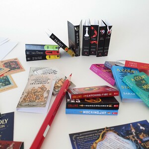 Peut inclure: Une collection de livres broch&eacute;s miniatures, dont des titres comme "Le Hobbit", "Les Deux Tours", "Une Cour d'&Eacute;pines et de Roses" et "Hunger Games". Les livres sont dispos&eacute;s sur une surface blanche avec un crayon rouge et d'autres articles li&eacute;s aux livres.