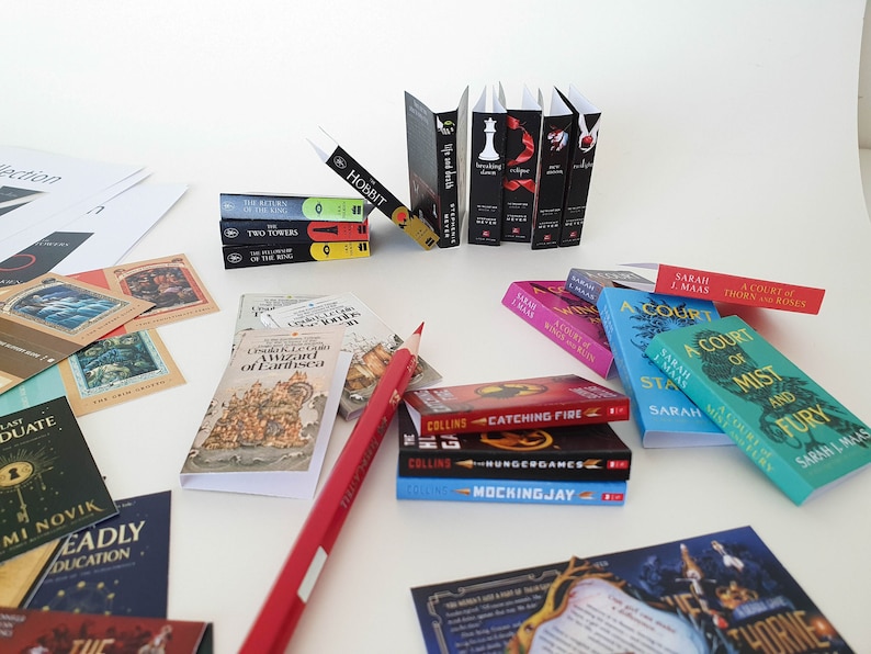 Peut inclure: Une collection de livres broch&eacute;s miniatures, dont des titres comme "Le Hobbit", "Les Deux Tours", "Une Cour d'&Eacute;pines et de Roses" et "Hunger Games". Les livres sont dispos&eacute;s sur une surface blanche avec un crayon rouge et d'autres articles li&eacute;s aux livres.